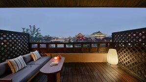 Un balcón con piso de madera, un sofá con cojines, una mesa con vela y vistas a edificios tradicionales.