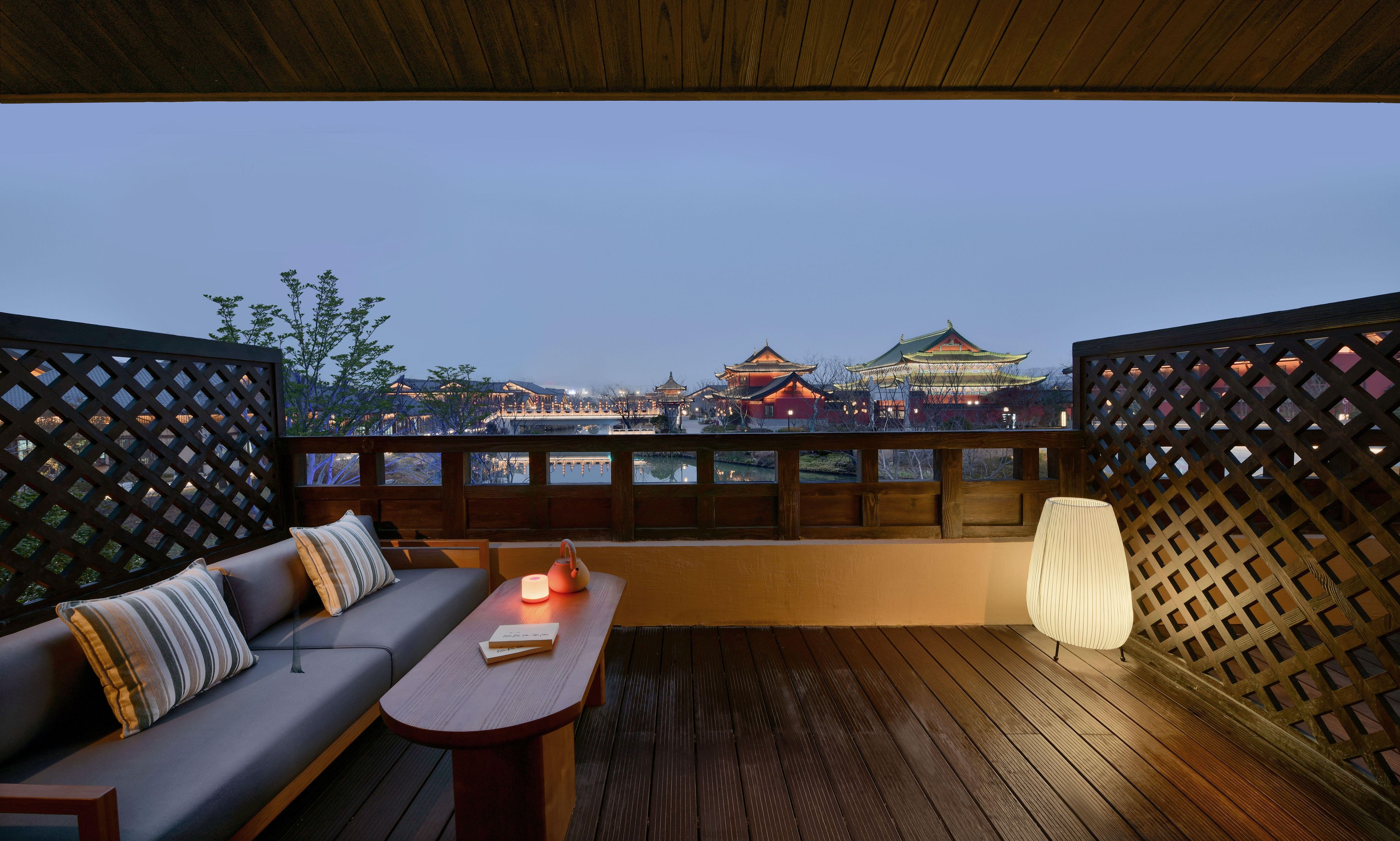 A balcony with a wooden floor, a sofa with cushions, a table with a candle, and a view of traditional buildings.