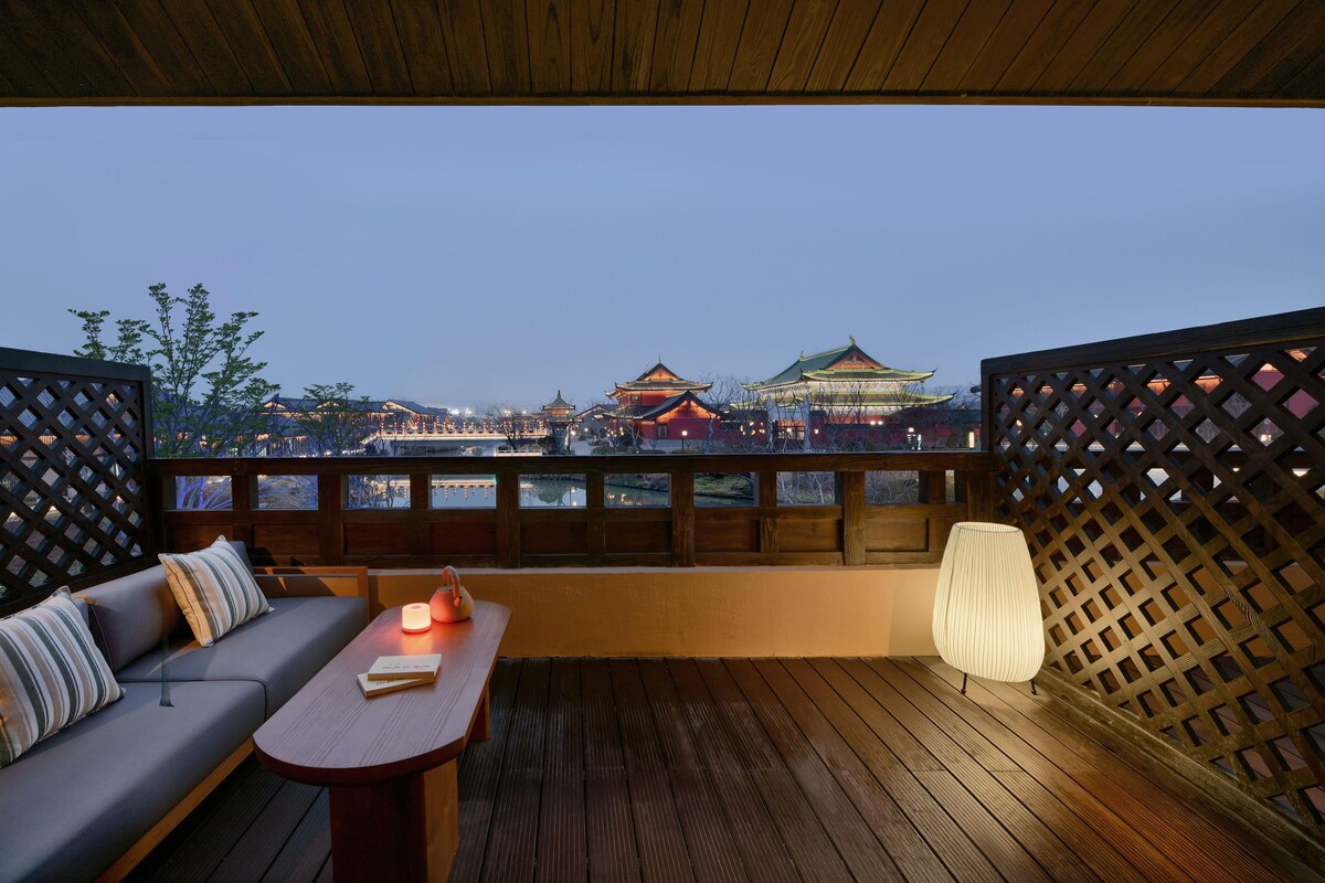 A balcony with a wooden floor, a sofa with cushions, a table with a candle, and a view of traditional buildings.
