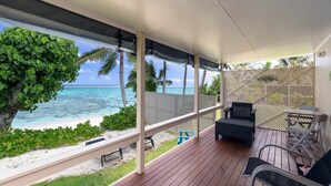 A covered outdoor deck with wooden flooring, a seating area with chairs and a table, and a view of the beach and ocean.