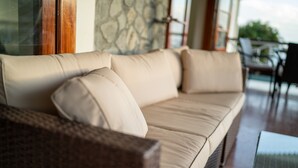 En beige sofa med puder, en fletstol og et bord med glasplade i et rum med stenvæg og udsigt ud