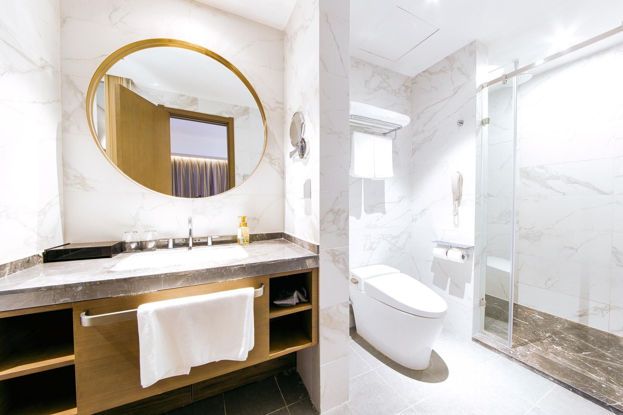 A modern bathroom with a marble vanity, a round mirror, a toilet, and a glass-enclosed shower.
