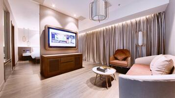 Ein modernes Hotelzimmer mit Flachbildfernseher an der Wand, einem Holzkabinett, einem runden Couchtisch, einer Couch und einem großen Fenster mit Vorhängen.