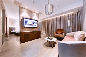 A modern hotel room with a flat-screen TV mounted on the wall, a wooden cabinet, a round coffee table, a sofa, and a large window with curtains.