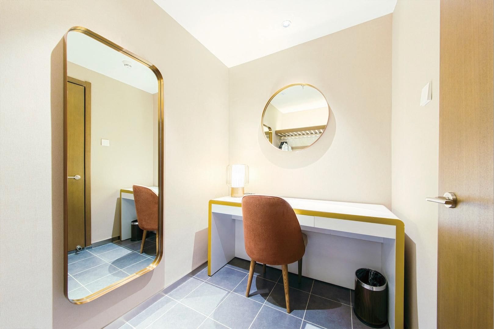 Un bagno d'albergo con uno specchio, uno specchio rotondo, una scrivania, una sedia e un cestino.