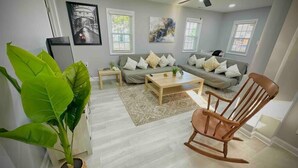 A modern living room with a sofa, coffee table, and a rocking chair. There are two windows with blinds, a ceiling fan, and a large green plant. - Colonial Beach Cottage, 7 minutes walk to beach! (Colonial Beach)