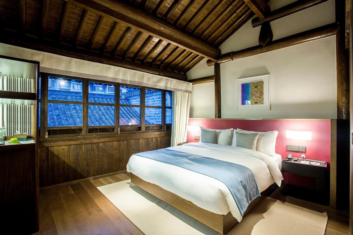 Quarto de hotel com uma cama grande, vigas de madeira no teto, uma janela com vista para um edifício azul e uma mesa de cabeceira com um abajur.