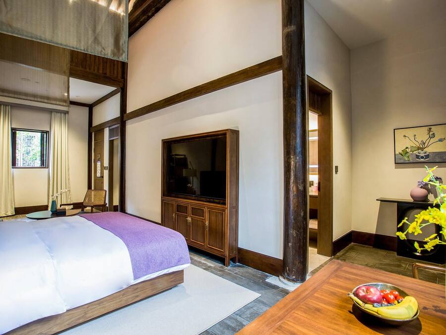 A hotel room with a bed, a television, a wooden cabinet, and a dining table with a fruit bowl.