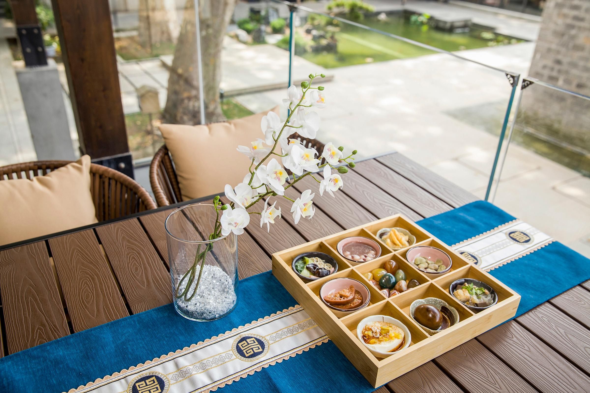 A wooden table with a glass vase of white flowers, a blue placemat, and a wooden tray with various food items.