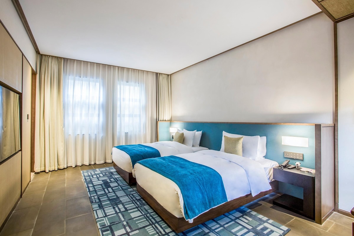 Habitación de hotel con dos camas, una mesita de noche, una lámpara y un ventanal con cortinas.