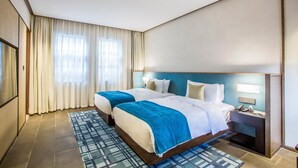 Habitación de hotel con dos camas, una mesita de noche, una lámpara y un ventanal con cortinas.