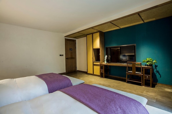 Quarto de hotel com duas camas, uma escrivaninha com cadeira, televisão e uma pequena mesa com planta.