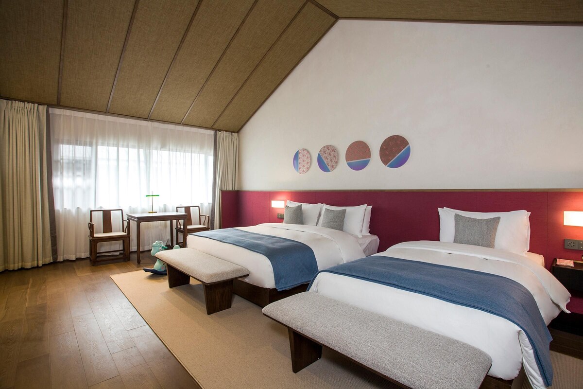 Quarto de hotel com duas camas, uma escrivaninha, uma cadeira e uma janela com cortinas.