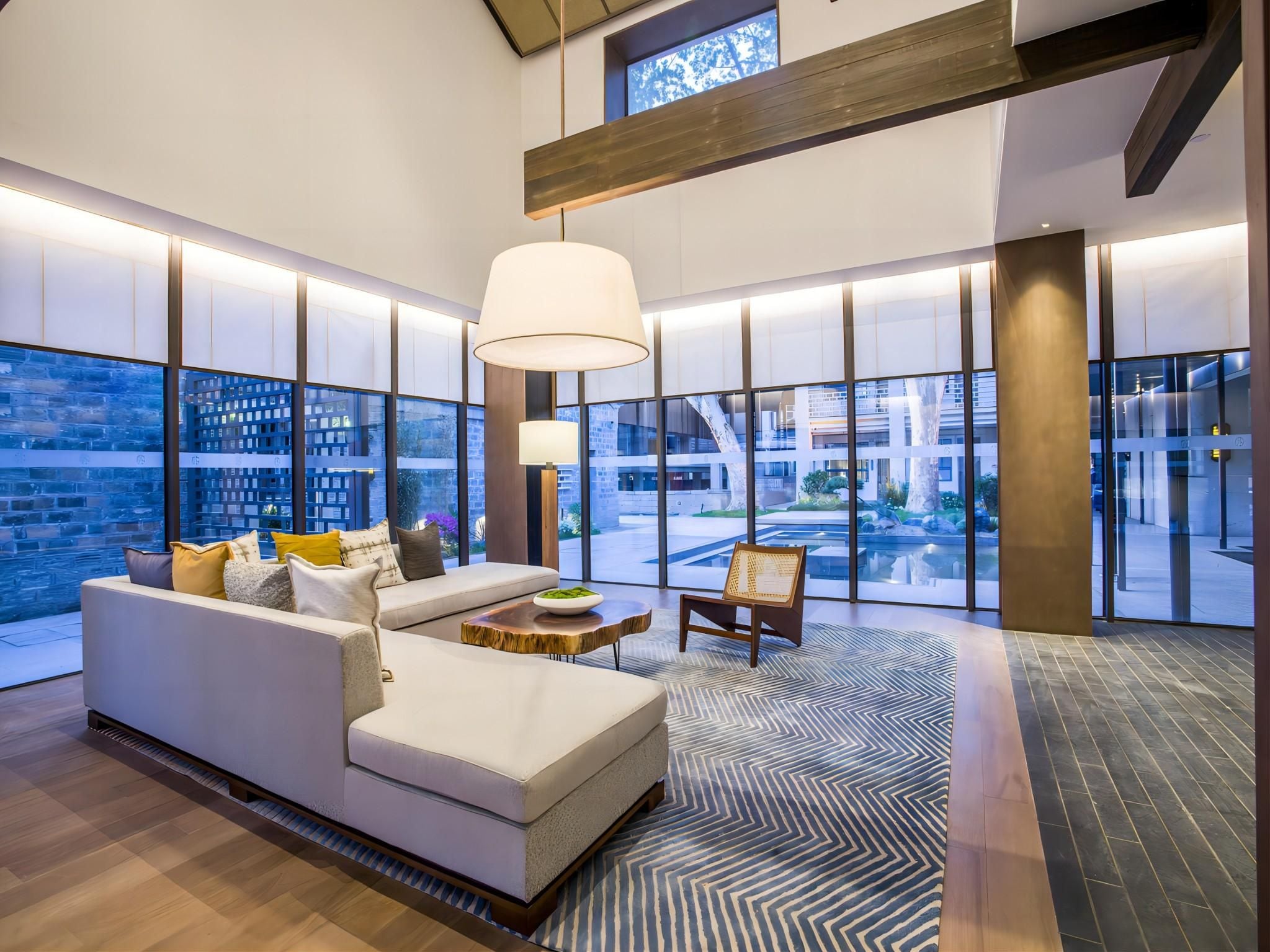 A modern hotel lobby with a large sofa, a coffee table, and a chair. There are large windows offering views of the outside.