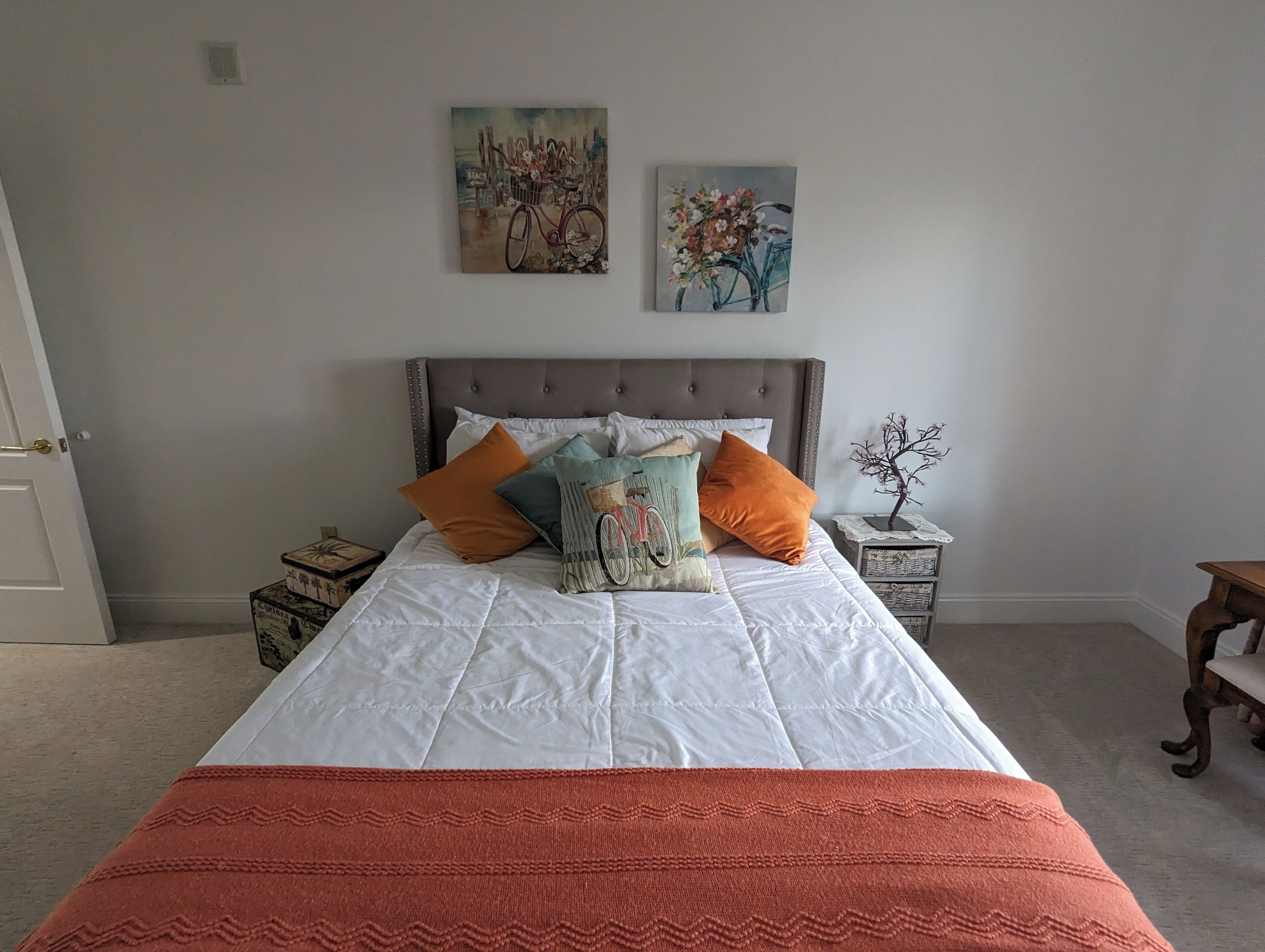 Ein ordentlich bezogenes Bett mit weißer Daunendecke und orangefarbener Tagesdecke, begleitet von zwei Nachttischen mit Dekoration.