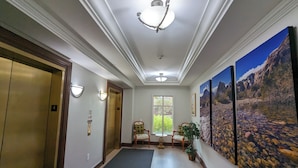 En korridor med två hissar, en sittgrupp med stolar, ett litet bord och en stor landskapsmålning på väggen.