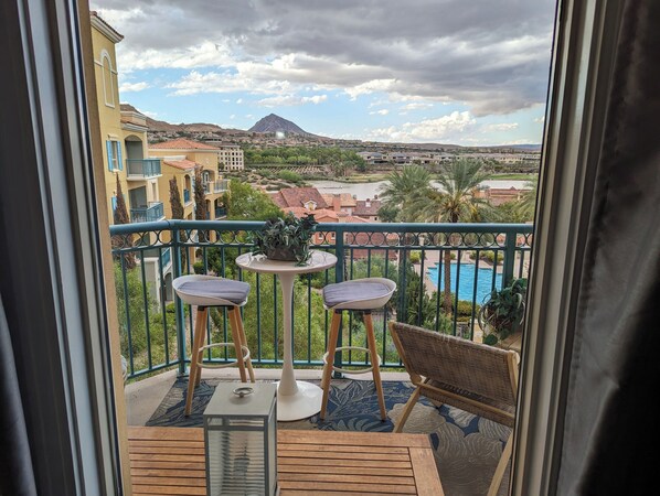 A balcony with two chairs and a small table, overlooking a pool and a mountainous landscape.