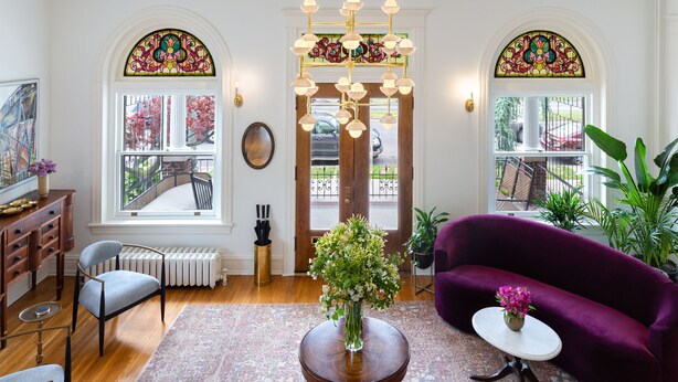A well-lit living room with a large window, a chandelier, a purple sofa, and a small table with a vase of flowers.