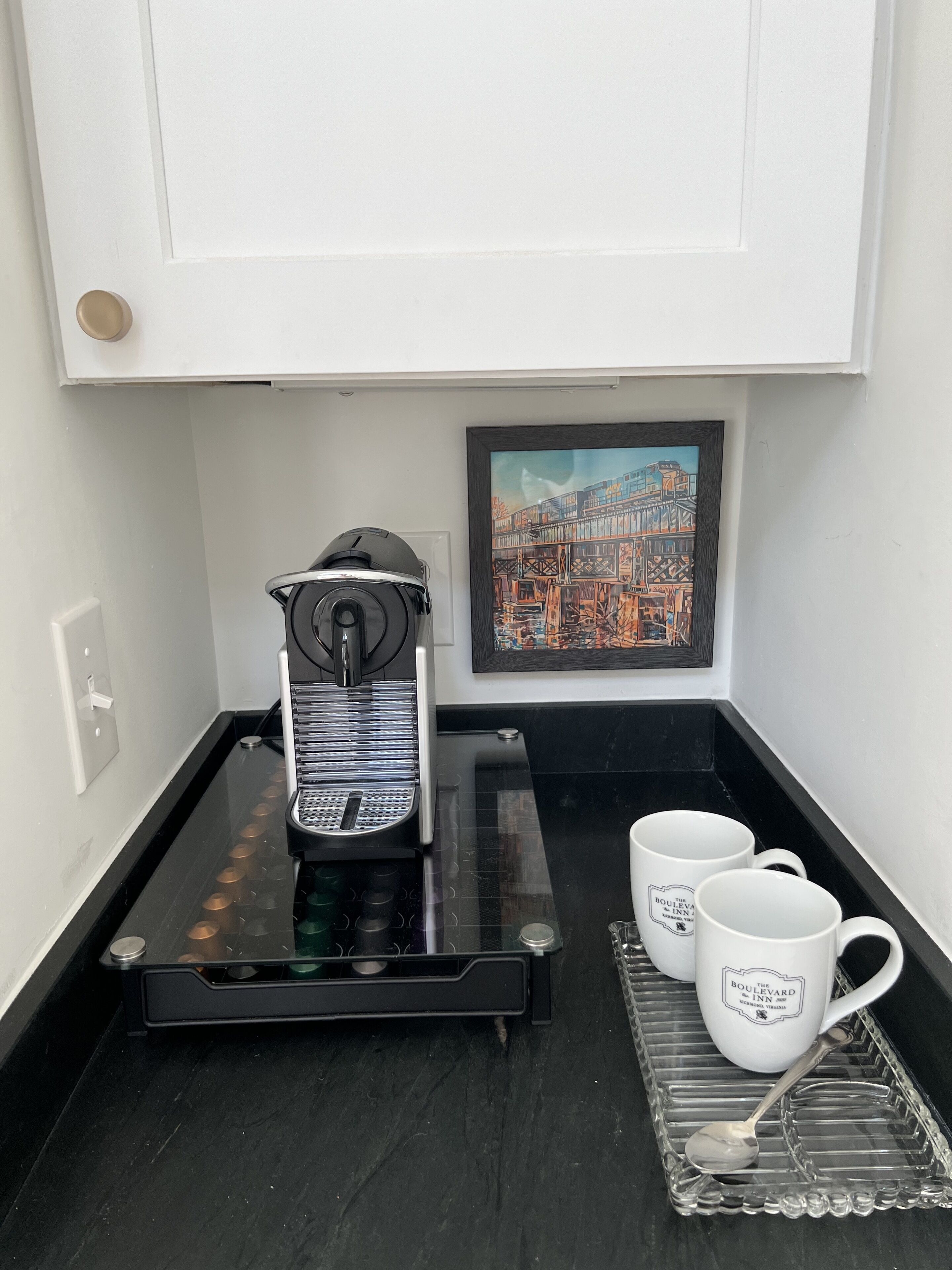 Una macchina per il caffè su un piano di lavoro nero, con due tazze bianche e un cucchiaio su un vassoio in vetro.