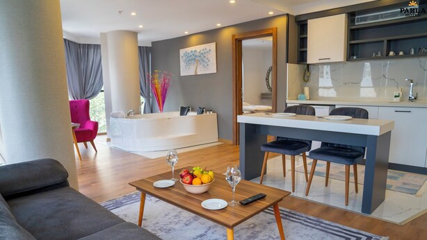 Una habitación de hotel moderna con sofá, una mesa de comedor con frutas y copas de vino, una zona de cocina y un espejo grande.