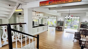 A spacious hotel lobby with a wooden floor, a staircase with white railings, a kitchen area, and a dining area with chairs and tables. - OCEAN Front Kona Studio - Spectacular view! (Kailua-Kona)