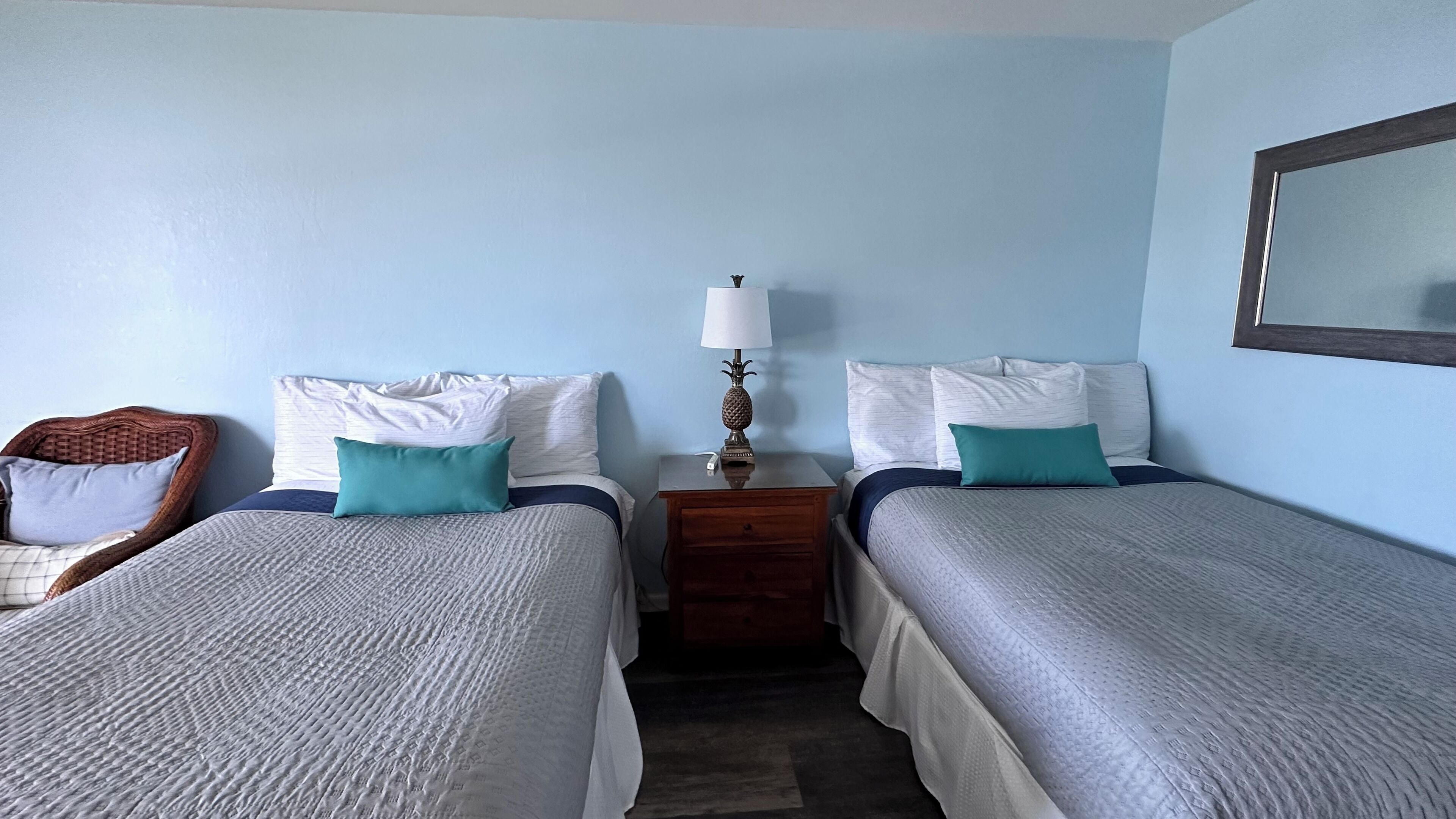 A hotel room with two single beds, a wooden nightstand with a lamp, a wicker chair, and a large mirror on the wall.