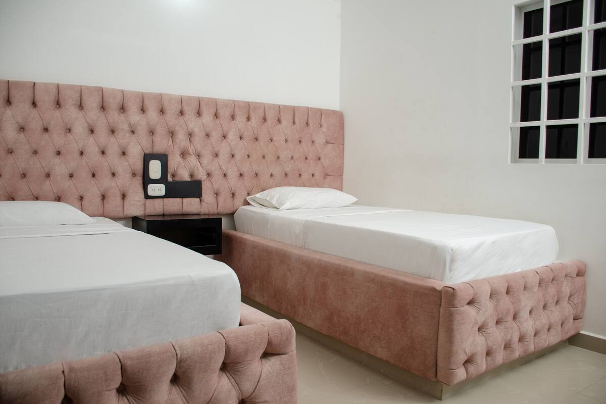 Une chambre avec deux lits, une tête de lit capitonnée, une table de chevet et une fenêtre avec des stores.
