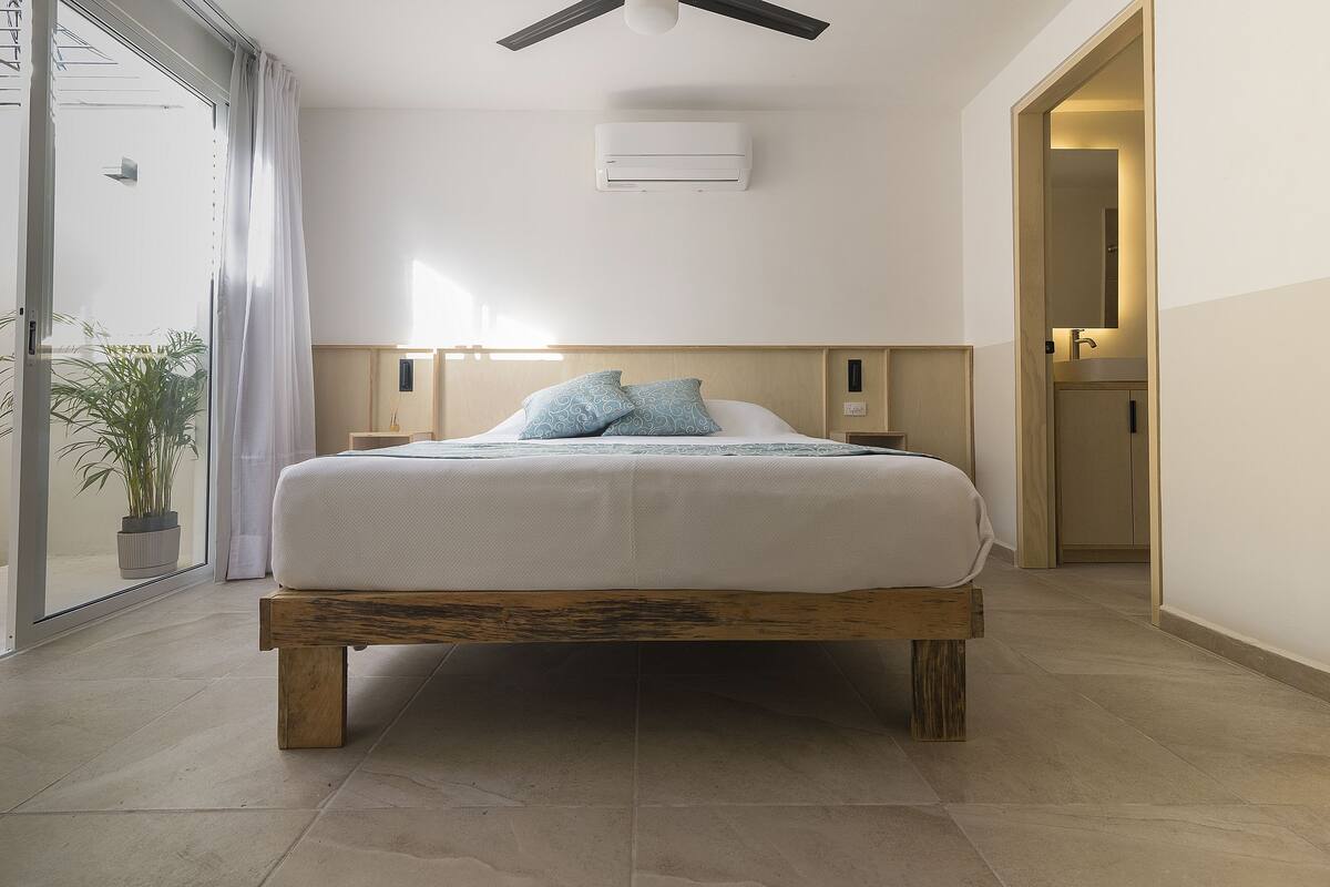 A modern bedroom with a wooden bed frame, a large window with curtains, a ceiling fan, and a small bedside table with a lamp.