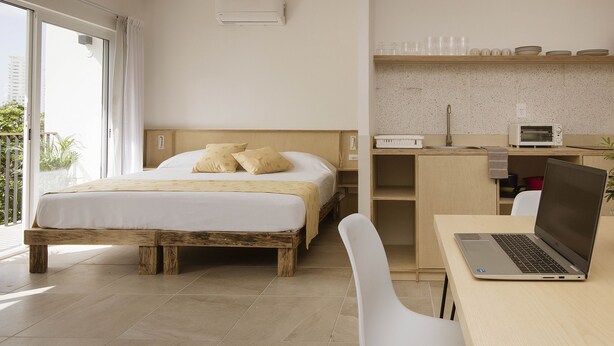 Un dormitorio con cama, escritorio con computadora portátil, kitchenette y puerta corrediza de vidrio al exterior.