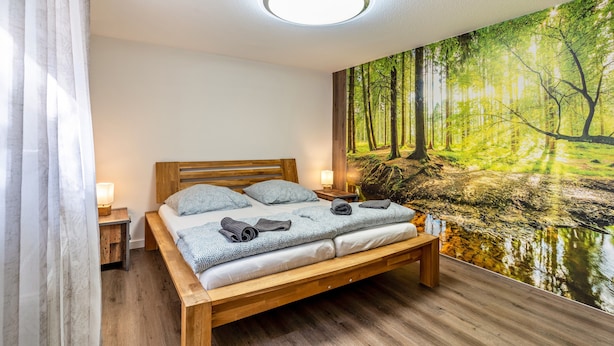 Un dormitorio con cama de madera, una mesita de noche, una lámpara y un mural de un bosque en la pared.