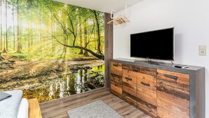 Una habitación moderna con un mural grande de un paisaje forestal, un televisor de pantalla plana, armarios de madera y un perchero.