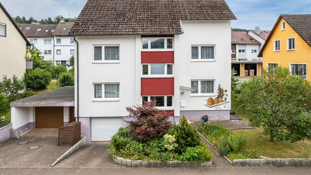 Ein mehrstöckiges Wohngebäude mit roter und weißer Fassade, einem kleinen Vorgarten und einem gepflasterten Einfahrt.