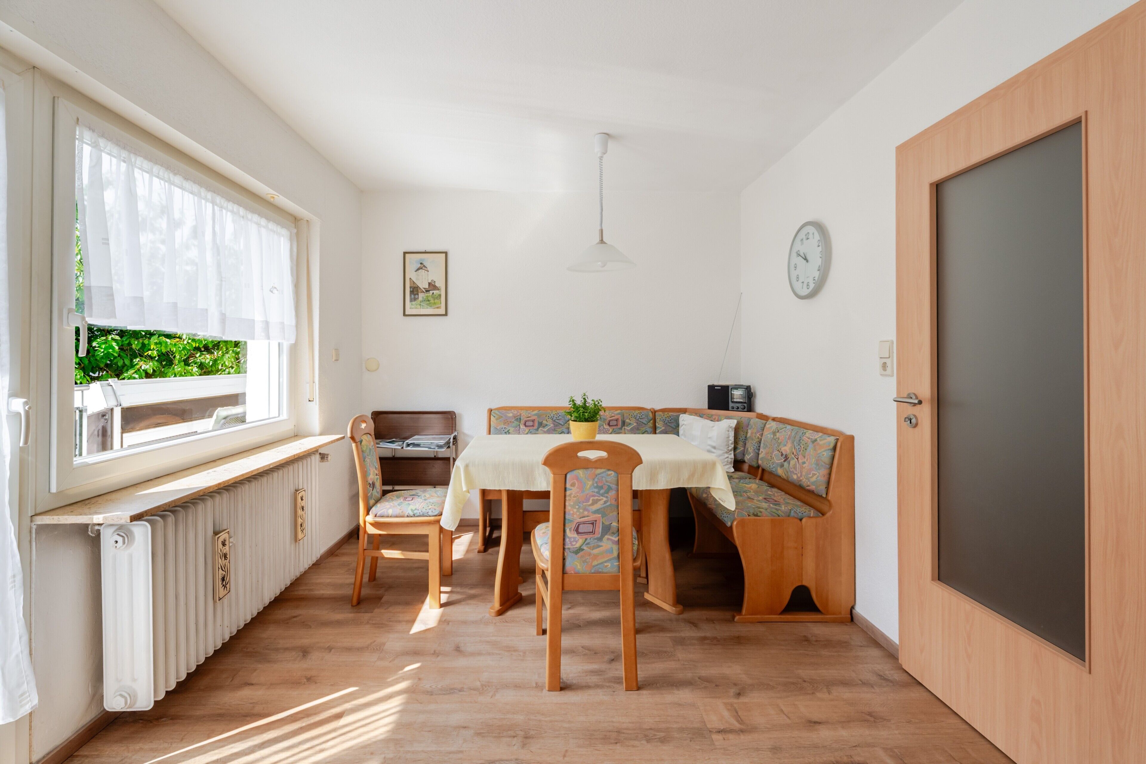 Ein Zimmer mit Esstisch, Stühlen, einer Bank, einem Heizkörper, einer Uhr und einer Tür.