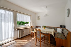 Une pièce comprenant une table à manger, des chaises, un canapé, un radiateur, une fenêtre avec des rideaux, une horloge murale et un luminaire suspendu.