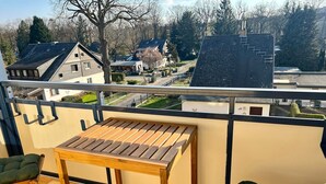 En balkon med et træbord og to grønne, polstrede stole, med udsigt over et boligområde med huse og træer.