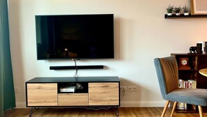En moderne stue med et fladskærms TV på væggen, et træunderholdningsmøbel med indbygget TV-reol, en grå polstret lænestol og en reol med forskellige genstande.