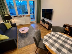 Un salon avec un canapé, une petite table ronde, une nappe à carreaux, un téléviseur et une fenêtre donnant sur des arbres.