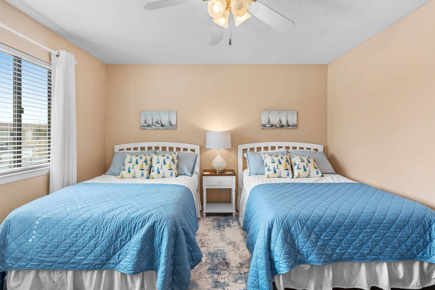 Une chambre avec deux lits, des murs de couleur beige, un ventilateur de plafond et une fenêtre avec des stores.