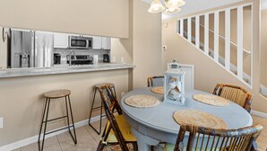 A compact kitchen with modern appliances, a dining area with a round table and wicker chairs, and a staircase with a white railing. - Sunray - Gulf-side townhouse, beach access & pool (Navarre)