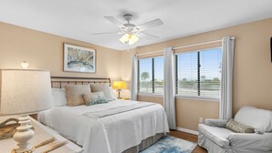 A bedroom with a bed, bedside table, lamp, and a view of the outdoors through large windows. - Sunray - Gulf-side townhouse, beach access & pool (Navarre)