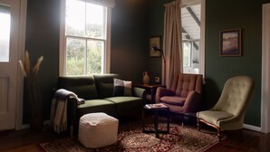 Een gezellige woonkamer met een groene bank, een bruine fauteuil en een witte poef. Op de vloer ligt een vloerkleed met een patroon en er staat een klein tafeltje met een lamp.