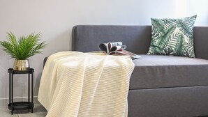 Een grijze bank met een witte gebreide plaid, een groen gestreept kussen, en een klein zwart bijzettafeltje met een potplant.