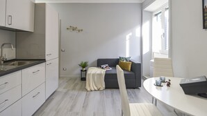 Een moderne keuken met witte kasten en een zwart aanrecht, een grijze bank met een geel kussen, een klein bijzettafeltje met een plant, en een eethoek met een witte tafel en stoelen.