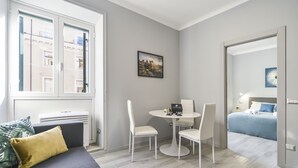 Een moderne hotelkamer met een bed, een eettafel met stoelen, een bank, een raam met uitzicht op gebouwen en een schilderij aan de muur.