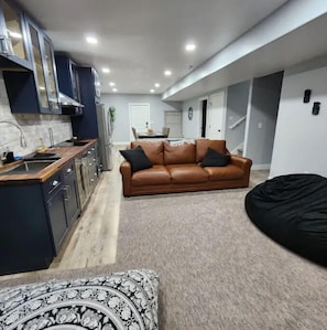 A modern kitchen with dark cabinets, a wooden countertop, and a built-in stove. There is a brown leather sofa with black cushions in the living area, and a black bean bag chair. - One Amazing Space (West Valley City)