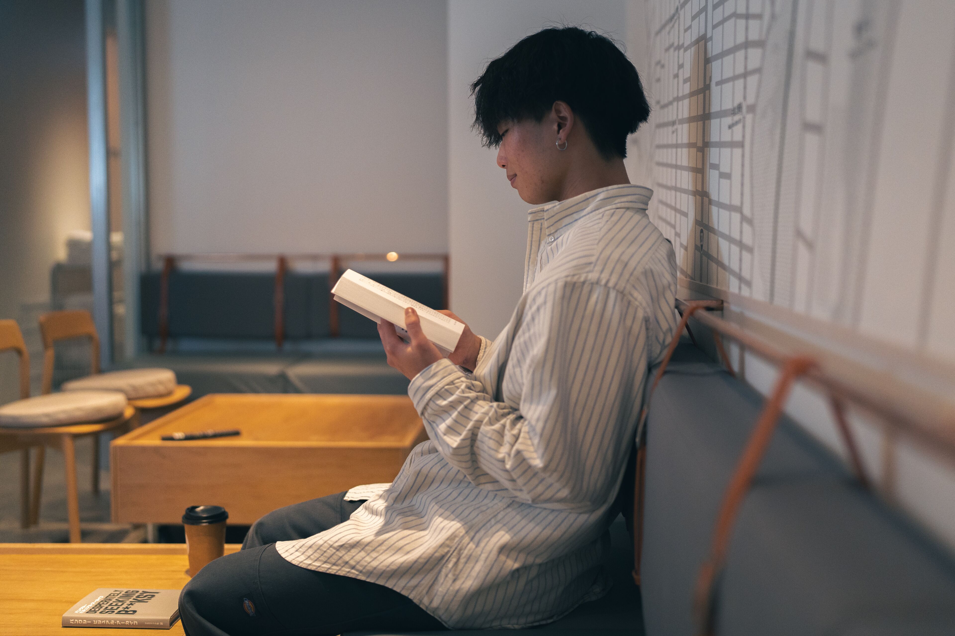 A person sitting at a wooden table in a cafe, reading a book. There are chairs and a wooden bench in the background.