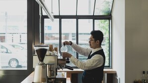Una persona manovra una macchina da caffè al bancone, con vista su auto e palazzi attraverso una finestra.