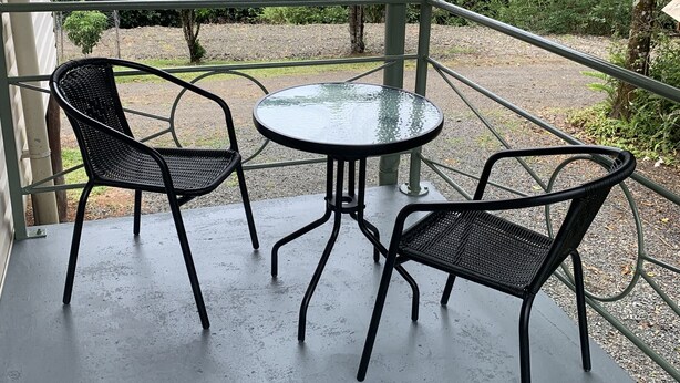 En overdækket terrasse med et rundt bord og to stole, omgivet af grøn vegetation og træer.