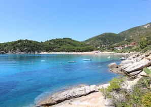 A coastal area with clear blue waters, rocky shores, and a hillside with buildings.