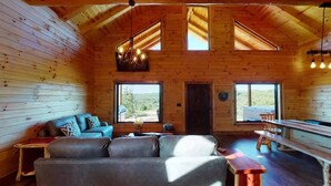 A wooden cabin interior with a sofa, a table, and a bench. There are two windows providing natural light and a view of the outdoors. - Stunning Hocking Hills Views, Huge Skies ~ Sauna, Pet Friendly, Hot Tub, WiFi (new plymouth)